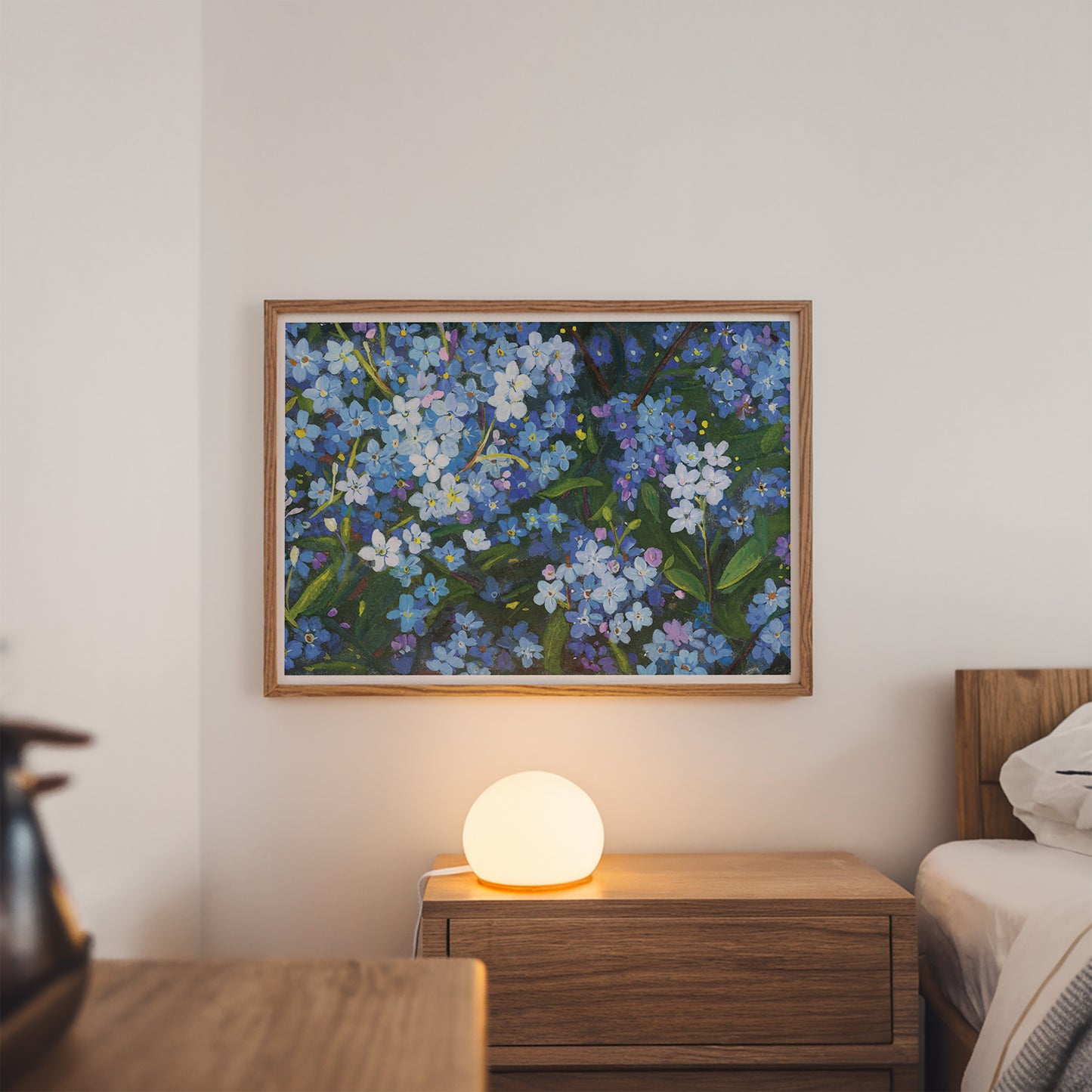 A framed floral painting featuring blue, purple, and white flowers with green leaves, hanging on a light beige wall above a wooden nightstand with a glowing spherical lamp, in a softly lit bedroom setting.