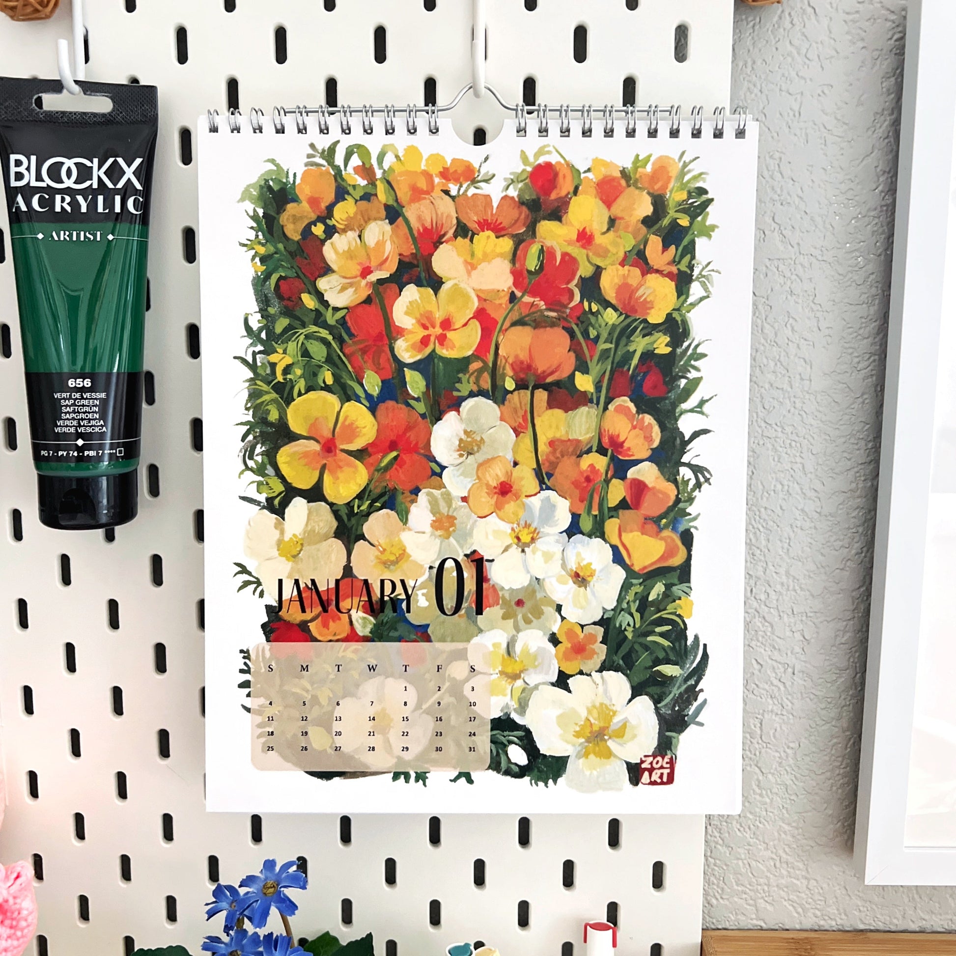Floral calendar hanging on a pegboard with Blockx acrylic paint tube and flowers below.