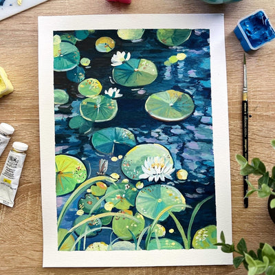 A watercolor painting of a pond with lily pads and white water lilies, displayed on a wooden desk surrounded by painting supplies including paint tubes, a palette, and brushes.