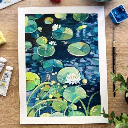 A watercolor painting of a pond with lily pads and white water lilies, displayed on a wooden desk surrounded by painting supplies including paint tubes, a palette, and brushes.