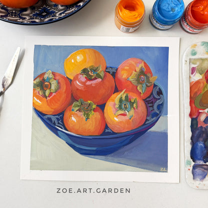 Framed artwork of persimmons in a blue bowl with painting supplies on a table.