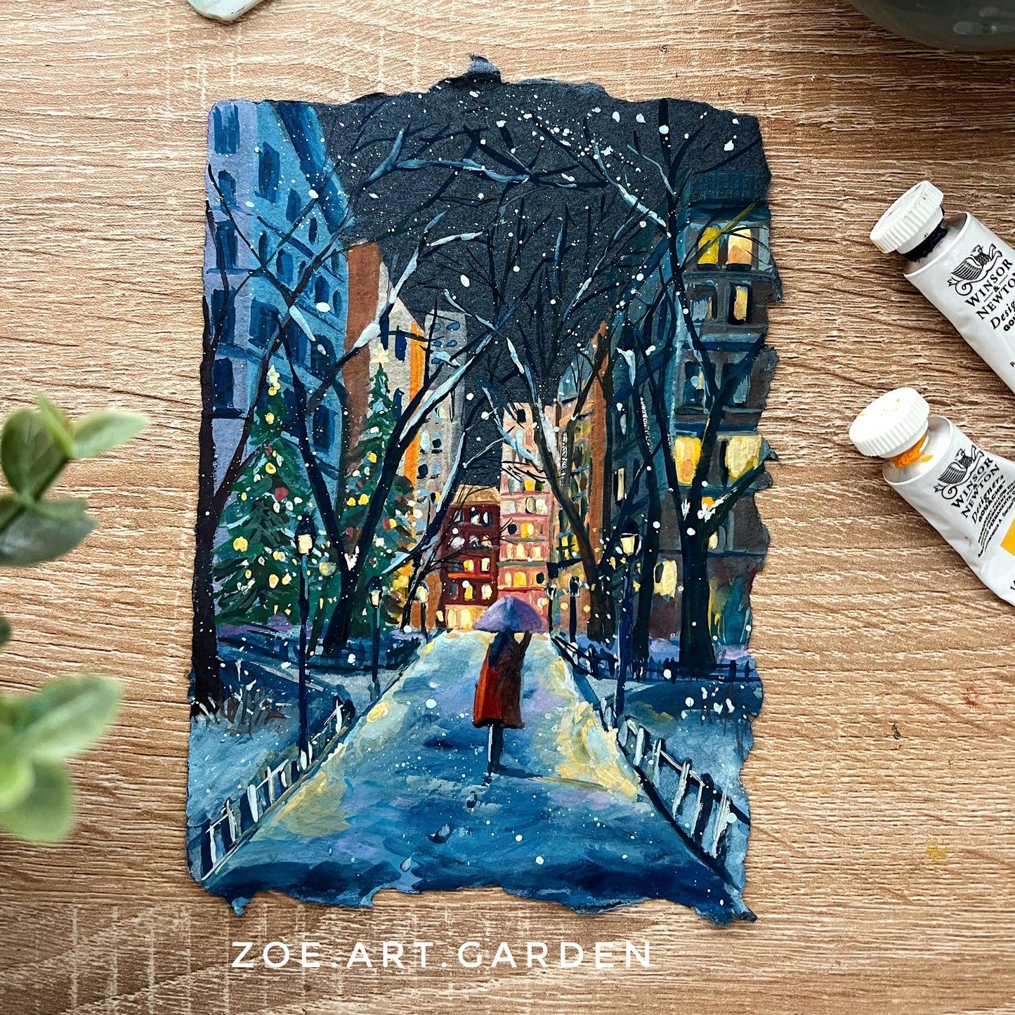Small painting of a snowy street scene with a person holding an umbrella, placed on a wooden surface with art supplies.