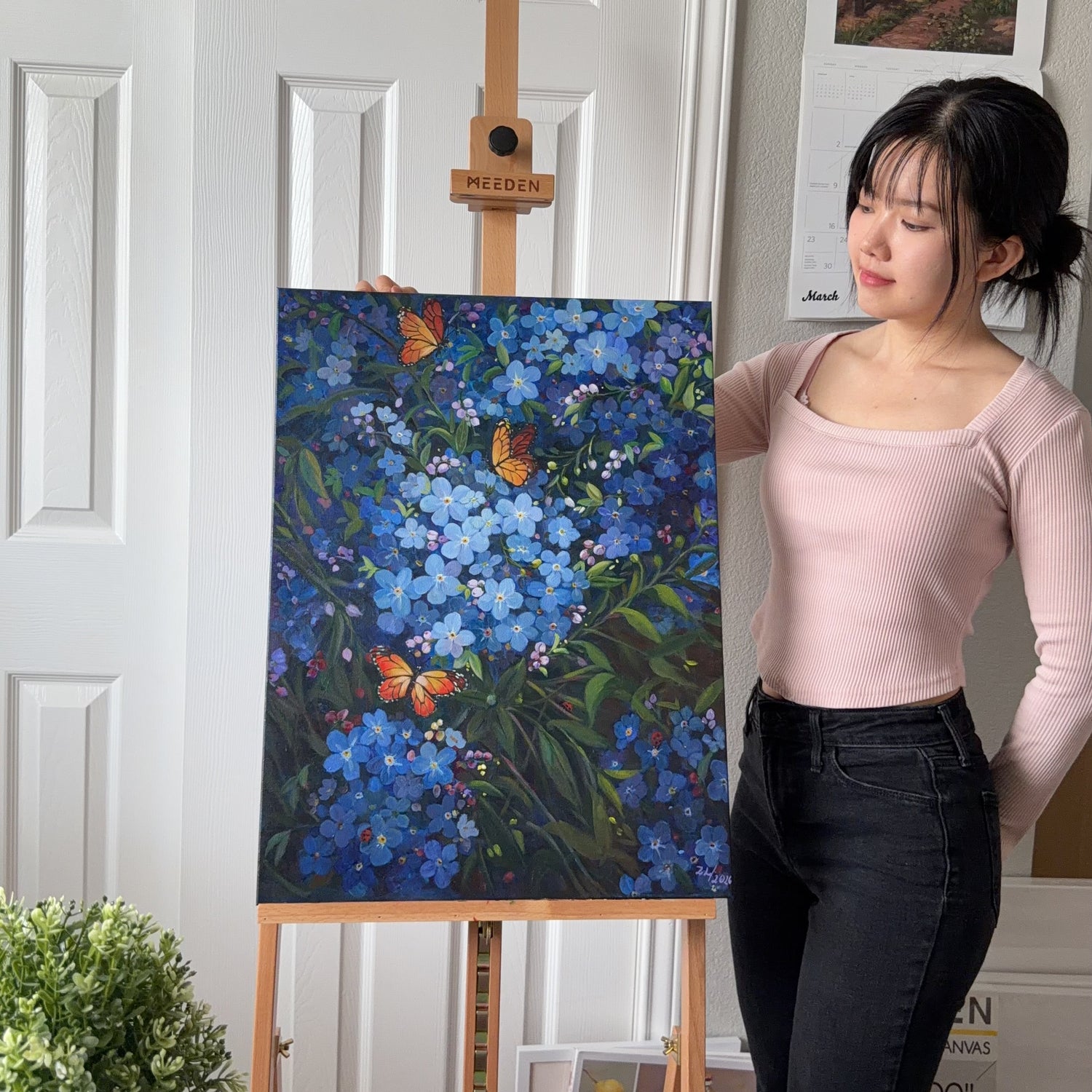 A woman stands beside a wooden easel holding a colorful painting of blue flowers and orange butterflies in front of a white paneled door.