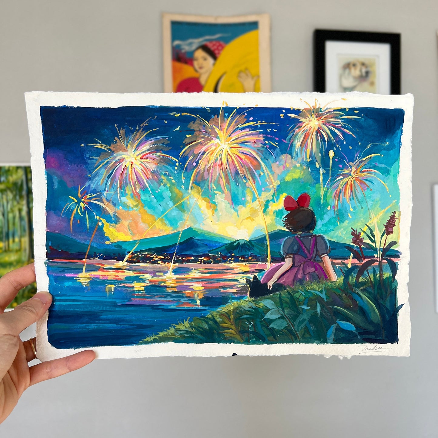 Hand holding a painting of a person watching fireworks over a body of water.