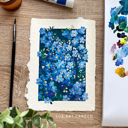 Artistic depiction of blue flowers on paper with painting supplies and a plant on a wooden surface.