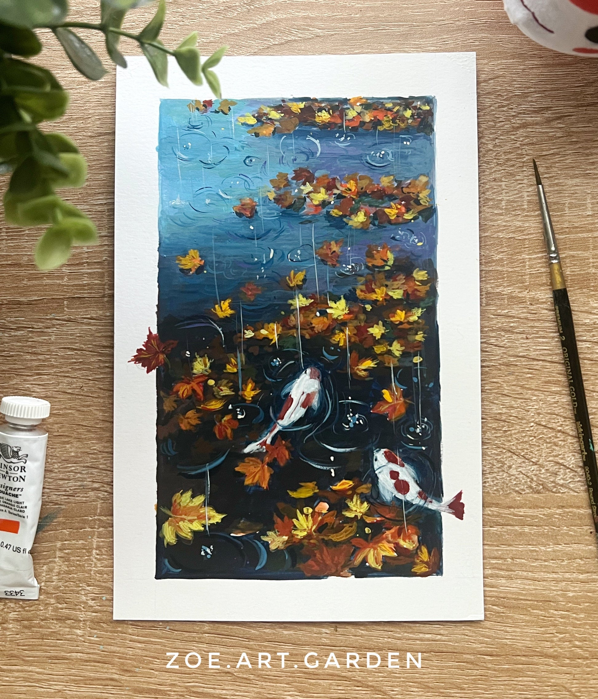 Art print of koi fish and autumn leaves on a wooden surface with a paint tube and brush.