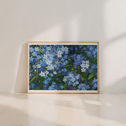 A framed floral art print featuring blue forget-me-nots with green leaves, displayed in a light wood frame against a warm off-white wall and floor.