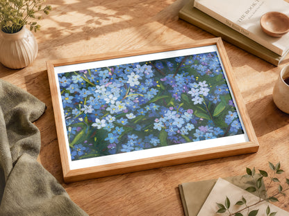 A framed floral art print featuring blue and white flowers with green leaves, displayed on a wooden table surrounded by books, a small plant, and a cup.