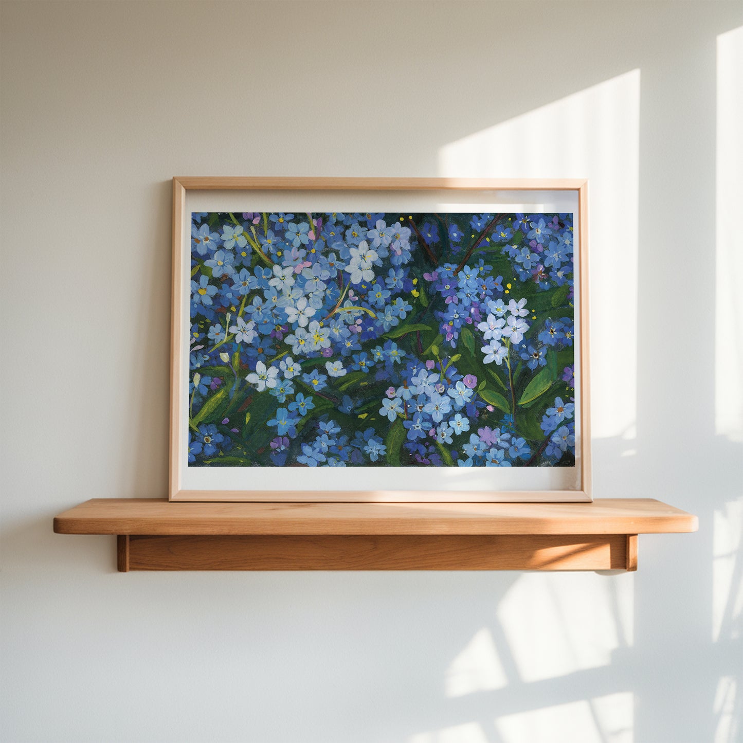 A framed floral painting of blue forget-me-nots with green leaves, displayed on a light wood floating shelf against a warm off-white wall with soft window shadows.