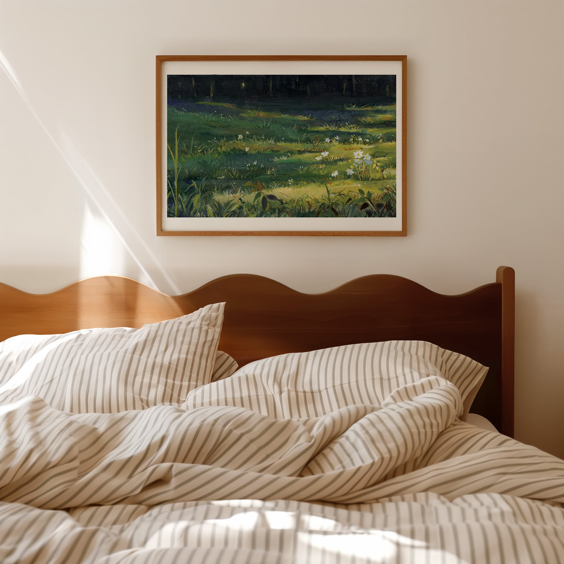 A framed landscape painting of a sunlit green meadow hangs on a beige wall above a wooden bed with cream and tan striped bedding.