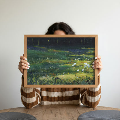 A person holds up a framed landscape painting of a grassy field with fireflies against a dark night sky. The person is wearing a brown and cream striped knit sweater and is seated at a wooden table against a plain light gray wall.