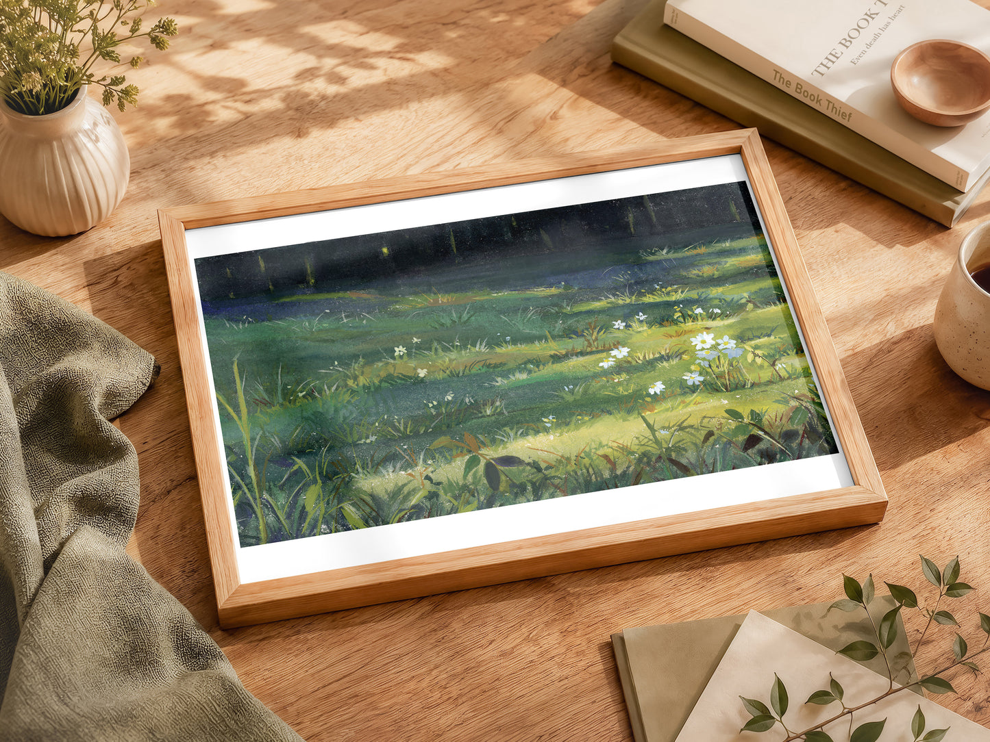 A framed landscape art print depicting a sunlit green meadow with wildflowers under a dark night sky, styled on a wooden table with books, a small plant, and a cup.