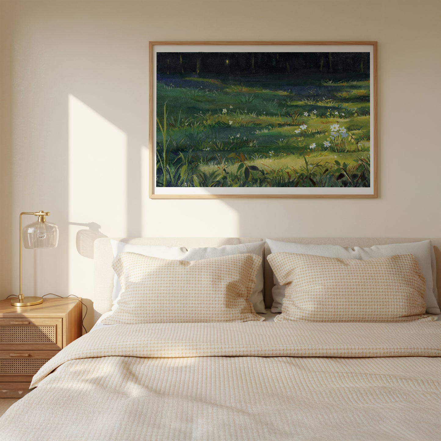 A framed landscape painting of a sunlit green meadow hangs on a beige wall above a neatly made bed with cream bedding and two patterned pillows. A wooden bedside table with a gold lamp sits to the left.