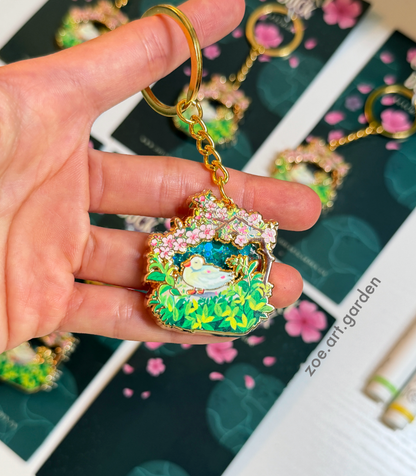 Hand holding a decorative keychain with floral design and bird illustration, with a blurred background featuring similar keychains.