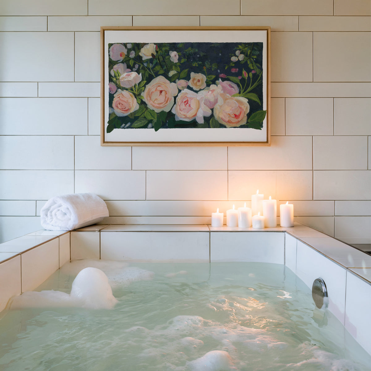 A framed floral painting of pink and white roses hangs on a white tiled wall above a bubble bath with lit candles on the ledge.