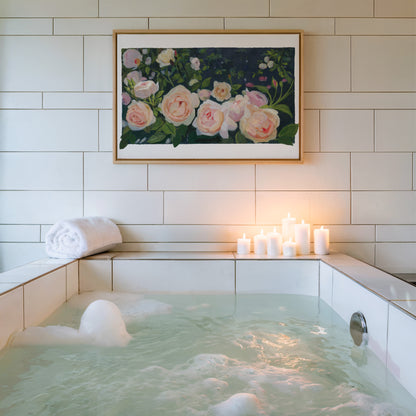 A framed floral painting of pink and white roses hangs on a white tiled wall above a bubble bath with lit candles on the ledge.
