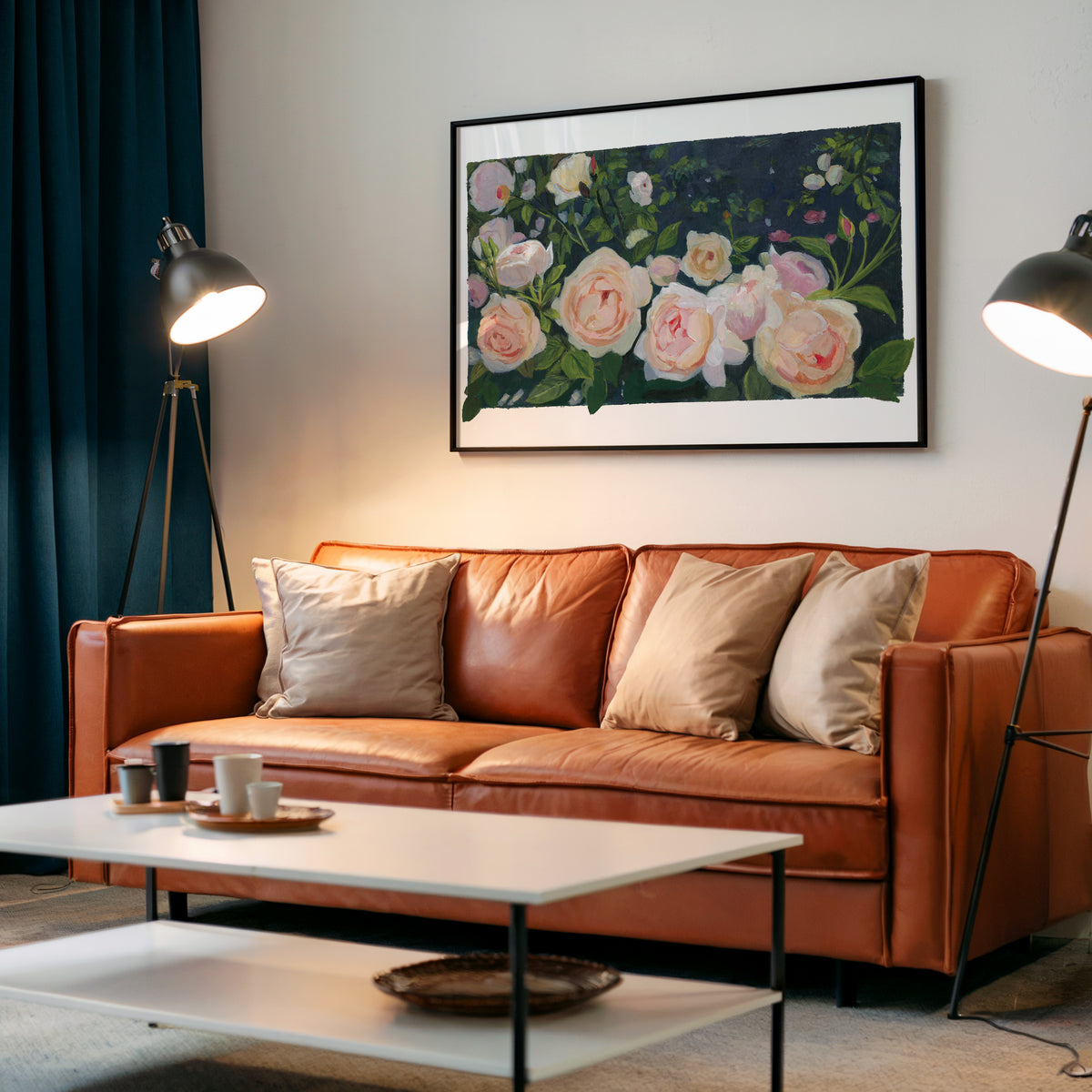 A framed floral painting hanging on a light gray wall above a rust-brown leather sofa in a modern living room setup with two floor lamps and a white coffee table.