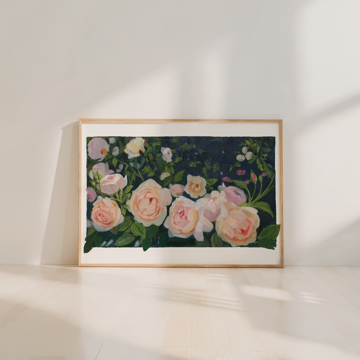 A framed floral painting of pale pink and white roses with green leaves, leaning against a white wall on a light wood floor. The artwork is enclosed in a thin light wood frame, and the scene is softly lit with natural shadows.