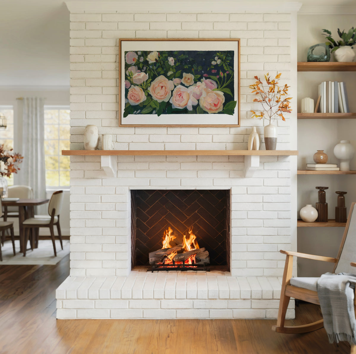 A framed floral painting hangs on a white brick fireplace mantel in a bright living room with a wood-burning fire, wooden flooring, and a rocking chair.