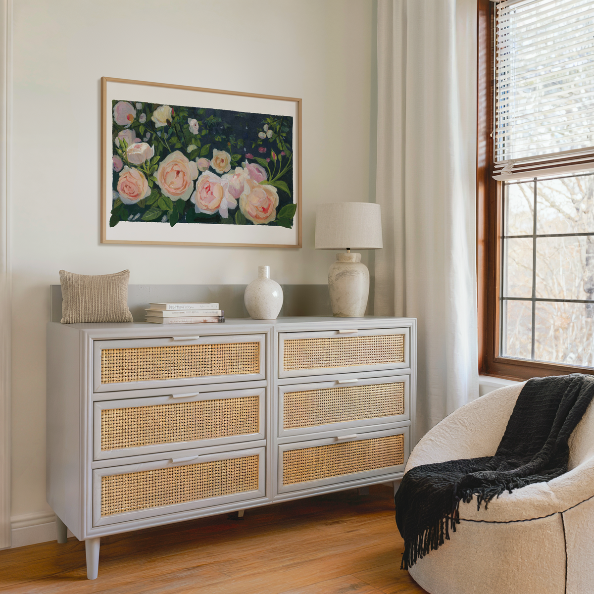 A light gray six-drawer dresser with woven cane panels in a styled room setting, decorated with a vase, books, and a small pillow, beneath a framed floral painting.