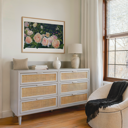A light gray six-drawer dresser with woven cane panels in a styled room setting, decorated with a vase, books, and a small pillow, beneath a framed floral painting.