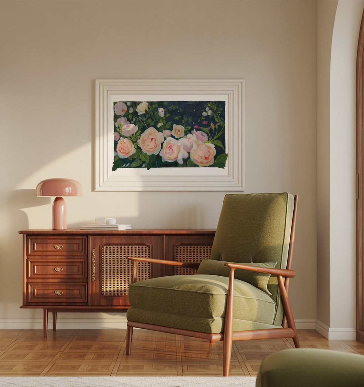 A framed floral painting hangs on a beige wall above a mid-century modern wooden credenza and olive green armchair. The painting features soft pink and white roses with green leaves on a dark background, enclosed in a white frame.
