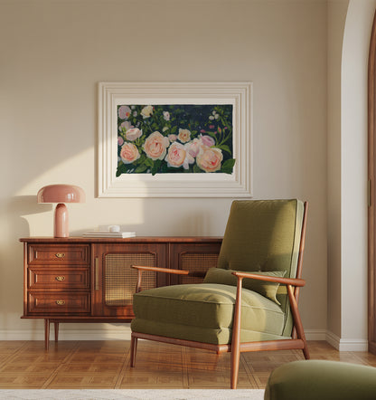 A framed floral painting hangs on a beige wall above a mid-century modern wooden credenza and olive green armchair. The painting features soft pink and white roses with green leaves on a dark background, enclosed in a white frame.