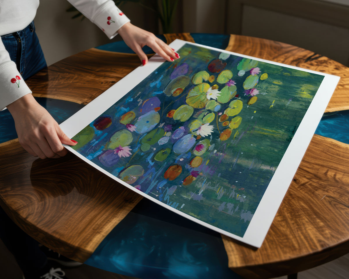 A person holds a large framed art print featuring an impressionistic pond scene with green and blue lily pads, pink flowers, and dark teal water, set on a polished wooden table.