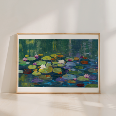 A framed painting of a pond with lily pads and water lilies in green, blue, yellow, pink, and white tones, leaning against a white wall on a light wood floor.