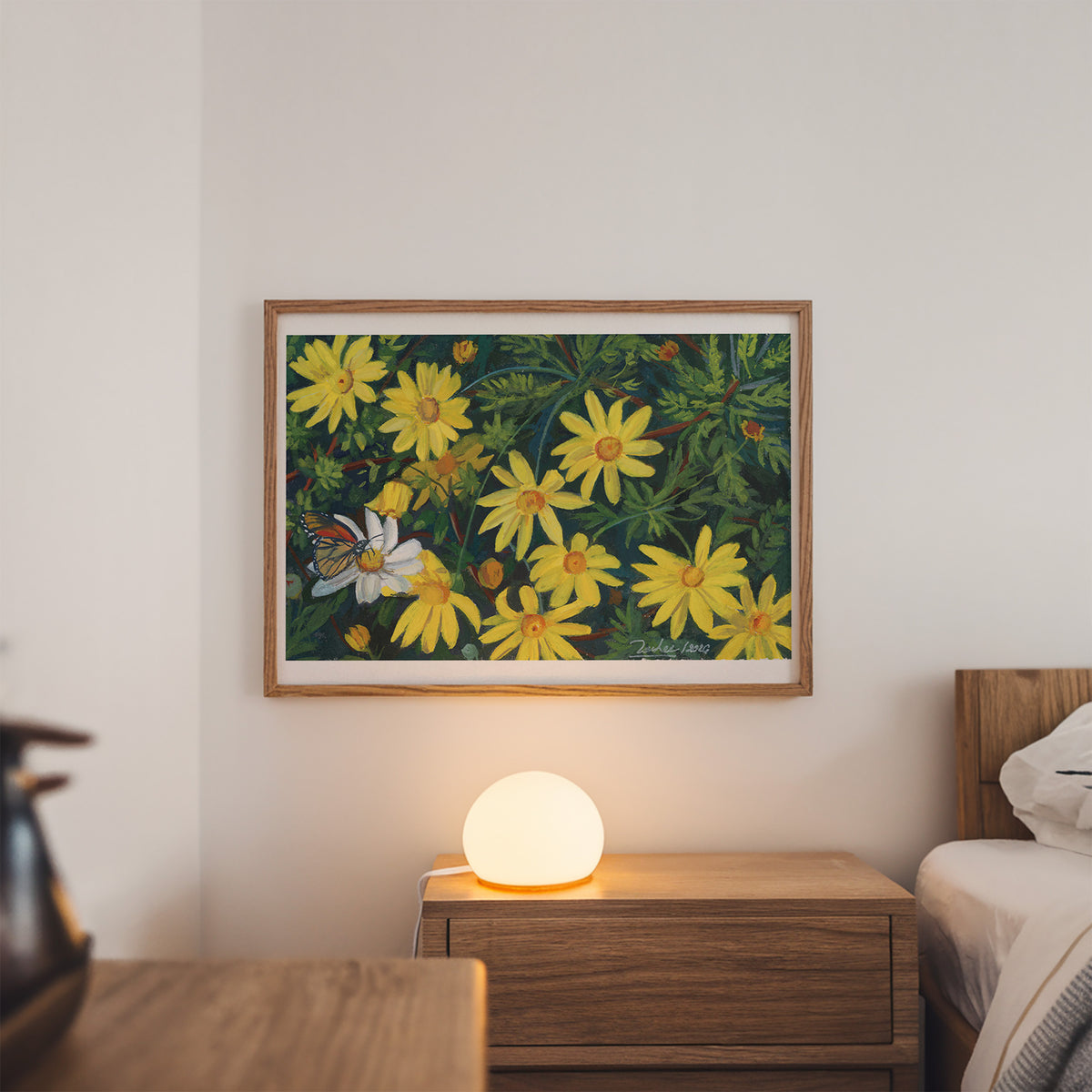 A framed floral art print featuring yellow flowers and a butterfly, displayed on a bedroom wall above a wooden nightstand with a glowing lamp.
